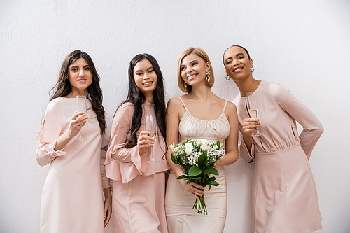 positivity, happy bride in wedding dress holding bridal bouquet and standing near interracial bridesmaids on grey background, champagne glasses, racial diversity, fashion, brunette and blonde women