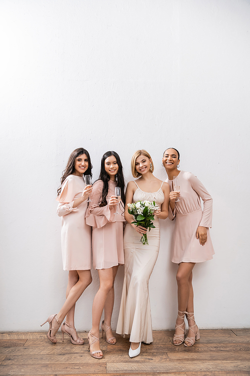 positivity, blonde bride in wedding dress holding bouquet and standing near interracial bridesmaids with champagne glasses on grey background, racial diversity, fashion, multicultural young women