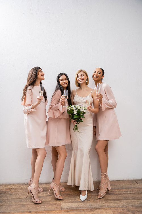 joy, blonde bride in wedding dress holding bouquet, standing with interracial bridesmaids, champagne glasses, grey background, racial diversity, fashion, multicultural young women