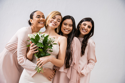 positivity, cheerful interracial bridesmaids hugging happy bride in wedding dress, bridal bouquet, grey background, racial diversity, fashion, brunette and blonde women, white flowers