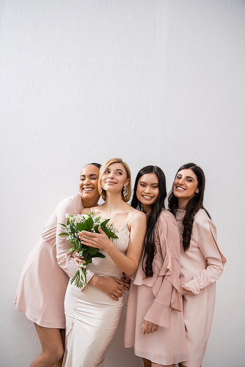 positivity, happy interracial bridesmaids hugging bride in wedding dress, bridal bouquet, grey background, racial diversity, fashion, brunette and blonde women, white flowers