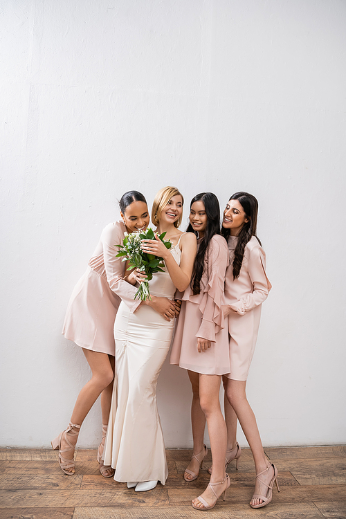 excitement, happy multicultural bridesmaids hugging bride in wedding dress, bridal bouquet, grey background, racial diversity, fashion, brunette and blonde, group of women, white flowers