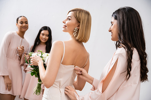 wedding preparations, bridesmaid zipping wedding dress of blonde bride, interracial women on blurred grey background, racial diversity, fashion, brunette and blonde women, bridal bouquet