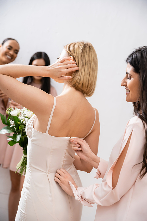 wedding preparations, happy bridesmaid zipping wedding dress of blonde bride, interracial women on blurred grey background, racial diversity, fashion, brunette and blonde women, bridal bouquet