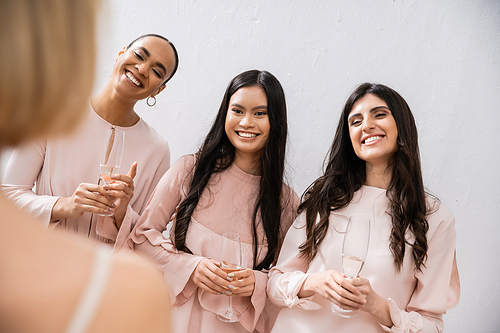 special occasion, wedding preparations, happy multicultural bridesmaids with glasses of champagne looking at bride on grey background, admire her style, fitting, bridesmaid gowns, diversity, blurred