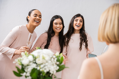 wedding preparations, cheerful multicultural bridesmaids with champagne looking at bride on grey background, admire her style, fitting, bridesmaid gowns, diversity, blurred