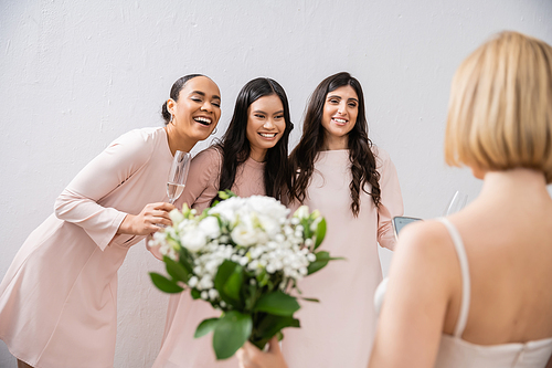 wedding preparations, cheerful multicultural bridesmaids with champagne looking at blonde bride on grey background, admire her style, fitting, bridesmaid gowns, diversity, blurred, special occasion