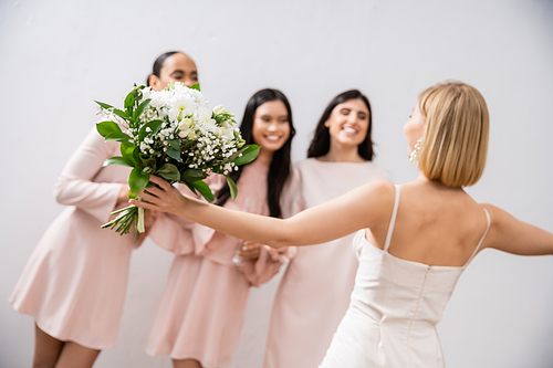 wedding preparations, cheerful bride with bouquet standing near blurred multicultural bridesmaids on grey background, dress fitting, bridesmaid gowns, wedding dress, diversity