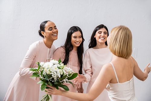 wedding preparations, cheerful multicultural bridesmaids with champagne looking at blonde bride with bouquet on grey background, admire her style, fitting, bridesmaid gowns, diversity