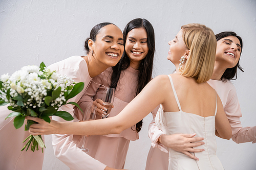 wedding preparations, happy bride with bouquet hugging cheerful multicultural bridesmaids with champagne on grey background, dress fitting, bridesmaid gowns, wedding dress, diversity