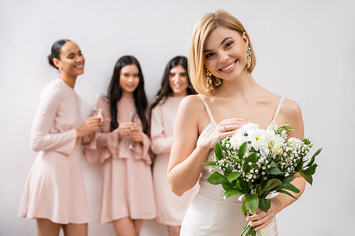 attractive bride in wedding dress holding bouquet, standing near blurred interracial bridesmaids on grey background, happiness, special occasion, blonde and brunette women