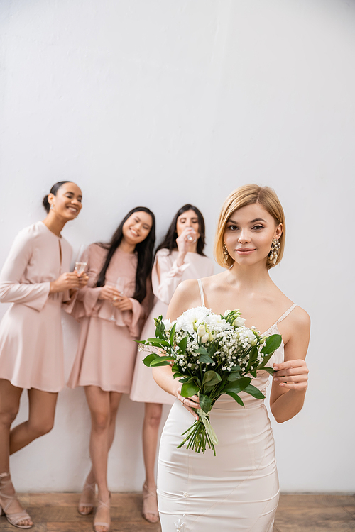 beautiful bride in wedding dress holding bridal bouquet, standing near blurred interracial bridesmaids on grey background, happiness, special occasion, blonde and brunette women