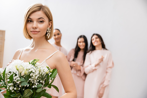 attractive bride in wedding dress holding bridal bouquet, standing near blurred interracial bridesmaids on grey background, happiness, special occasion, blonde and brunette women