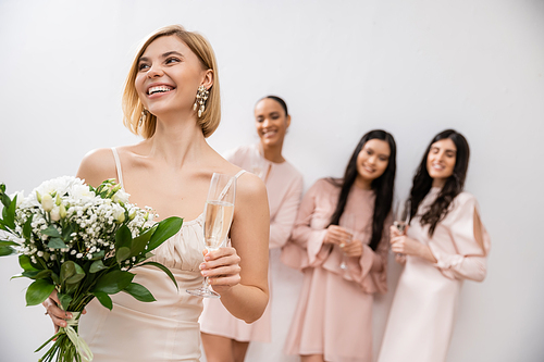 cheerful bride in wedding dress holding bridal bouquet and champagne glass, standing near blurred interracial bridesmaids on grey background, happiness, special occasion, blonde and brunette women