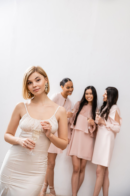 beautiful bride in wedding dress holding glass of champagne, standing near blurred interracial bridesmaids on grey background, happiness, special occasion, blonde and brunette women