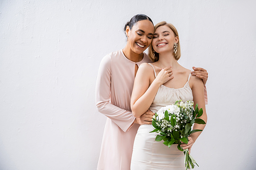 special occasion, happy bride with bridesmaid, positive interracial women, wedding dress and bridesmaid gown, african american woman hugging engaged friend on grey background