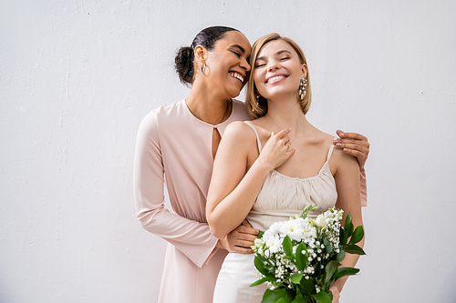 special occasion, cheerful bride with bridesmaid, happy interracial women, wedding dress and bridesmaid gown, african american woman hugging engaged friend on grey background