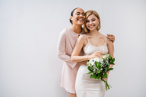 bridal shower, happy bride with bridesmaid, cheerful interracial women, wedding dress and bridesmaid gown, positive african american woman hugging engaged friend on grey background