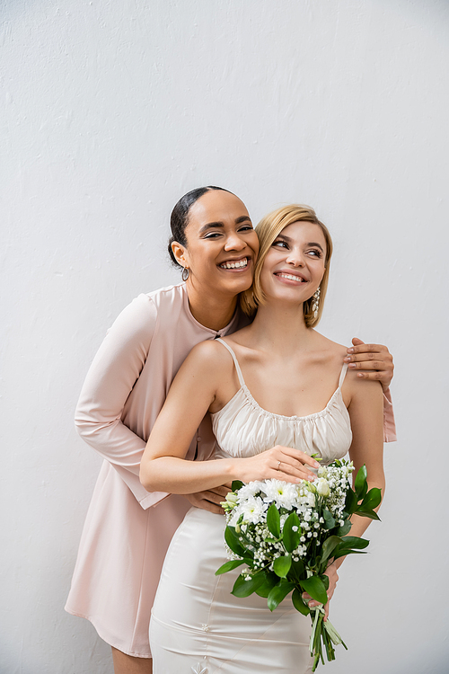 bridal party, happy bride with bridesmaid, cheerful interracial women, wedding dress and bridesmaid gown, african american woman hugging engaged friend on grey background