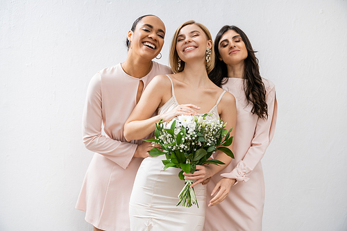 bridal party, special occasion, brunette and interracial bridesmaids hugging blonde bride, friendship goals, grey background, happy multicultural girlfriends, cultural diversity, bridal bouquet