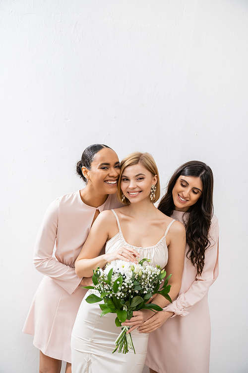 bridal party, special occasion, brunette and interracial bridesmaids hugging blonde bride, friendship goals, grey background, cheerful multicultural girlfriends, cultural diversity, bridal bouquet