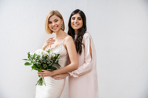 special occasion, cheerful bridesmaid hugging bride, friendship goals, grey background, happy girlfriends, bridal bouquet, blonde and brunette women, white flowers, happiness