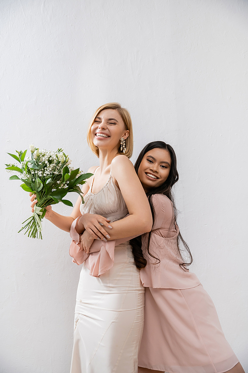 special occasion, asian bridesmaid hugging beautiful bride, friendship goals, grey background, happy girlfriends, bridal bouquet, blonde and brunette women, white flowers, happiness