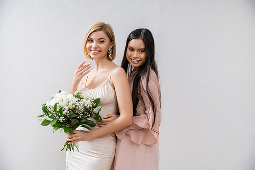 special occasion, asian bridesmaid hugging beautiful bride, friendship goals, grey background, happy girlfriends, bridal bouquet, blonde and brunette women, white flowers, positivity