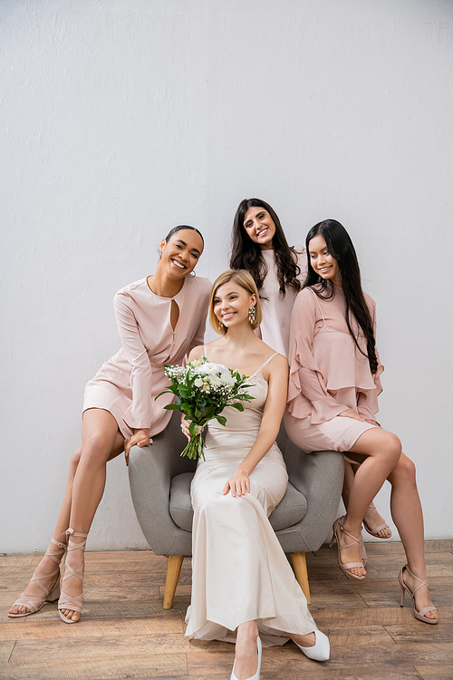 wedding photography, four women, bride and bridesmaids, interracial girlfriends, wedding day, cultural diversity, sitting on armchair, grey background, happiness and joy, bridal gown