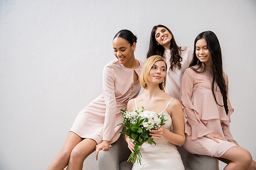 wedding photography, four women, joyful bride and bridesmaids, interracial girlfriends, wedding day, cultural diversity, sitting on armchair, grey background, happiness and joy, bridal gown