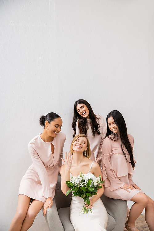 wedding photography, diversity, four women, joyful bride with bouquet showing her engagement ring near bridesmaids, wedding day, sitting on armchair, grey background, happiness and joy