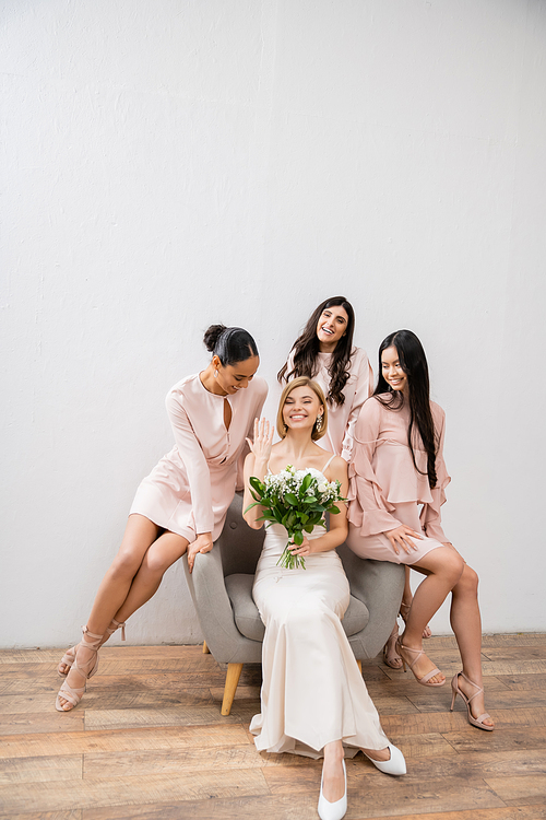 wedding photography, cultural diversity, four women, joyful bride with bouquet showing her engagement ring near bridesmaids, wedding day, sitting on armchair, grey background, happiness and joy