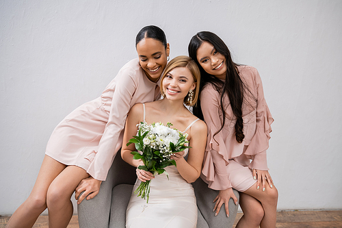 wedding photography, diversity, three women, happy bride with bouquet and her interracial bridesmaids sitting on armchair on grey background, brunette and blonde, joy, celebration