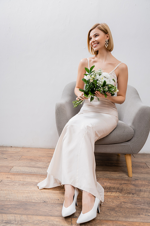 wedding photography, special occasion, beautiful, blonde bride in wedding dress sitting in armchair and holding bouquet on grey background, white flowers, bridal accessories, happiness