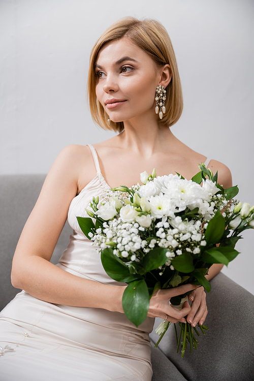 special occasion, beautiful and blonde bride in wedding dress sitting in armchair and holding bouquet on grey background, white flowers, bridal accessories, feminine, blissful