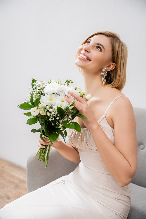 attractive and blonde bride in wedding dress sitting in armchair and holding bouquet on grey background, white flowers, bridal accessories, happiness, special occasion, smiling, feminine, blissful