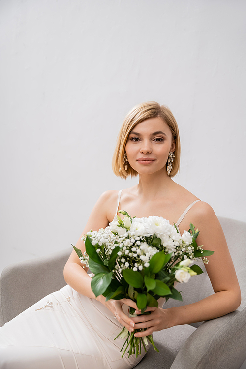 elegant and blonde bride in wedding dress sitting in armchair and holding bouquet on grey background, white flowers, bridal accessories, happiness, special occasion, beautiful, feminine, blissful