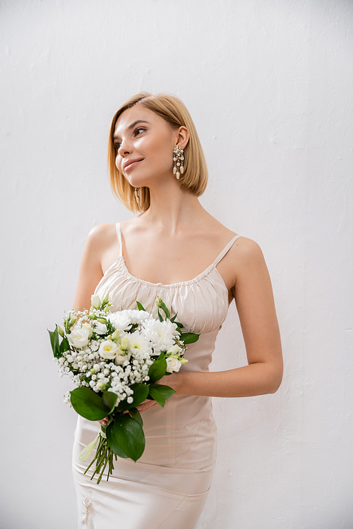 attractive and blonde bride in wedding dress holding bouquet on grey background, white flowers, bridal accessories, happiness, special occasion,   beautiful, feminine, blissful