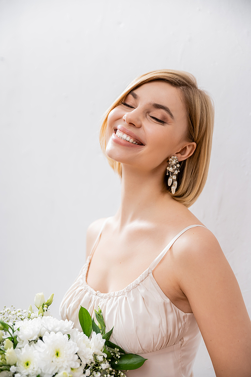 beautiful and blonde bride in wedding dress holding bouquet on grey background, joy, white flowers, bridal accessories, happiness, special occasion, feminine, blissful