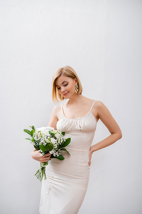 happy and blonde bride in wedding dress holding bouquet on grey background, white flowers, bridal accessories, happiness, special occasion,   beautiful, feminine, blissful