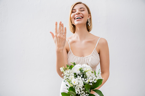 special occasion, gorgeous blonde bride in wedding dress holding bouquet and showing engagement ring, white flowers, bridal accessories, happiness, grey background