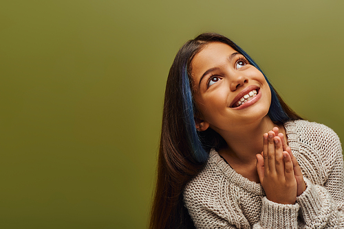 Portrait of smiling brunette preteen girl with dyed hair wearing knitted sweater while doing praying hands gesture and standing isolated on green, fashion-forward preteen with sense of style