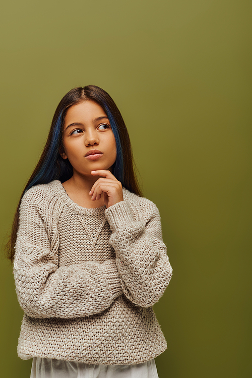 Pensive brunette preteen girl with dyed hair wearing stylish knitted sweater and looking away while standing isolated on green, fashion-forward preteen with sense of style