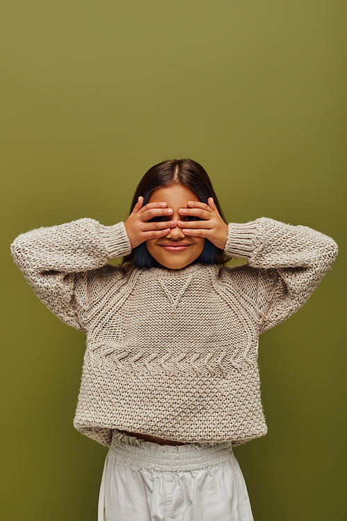 Smiling preteen child with colored hair wearing stylish knitted sweater while covering face with hands and standing isolated on green, fashion-forward preteen with sense of style