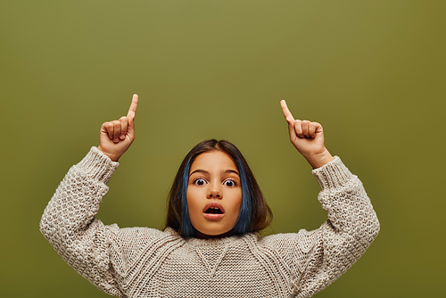 Shocked preteen girl with colored hair wearing stylish knitted sweater while pointing with fingers and looking at camera isolated on green, fashion-forward preteen with sense of style