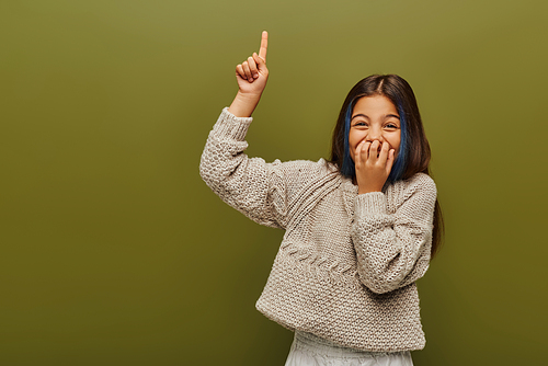 Laughing and stylish preteen girl with dyed hair wearing knitted sweater and pointing with finger while standing and posing isolated on green, fashion-forward preteen with sense of style