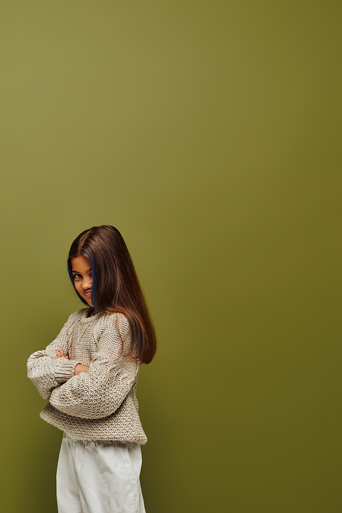 Offended and stylish preteen girl with colored hair wearing knitted sweater and crossing arms while standing and posing isolated on green, fashion-forward preteen with sense of style