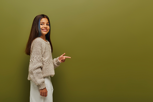 Smiling brunette preteen girl with dyed hair wearing trendy knitted sweater and pointing with finger while looking at camera on green background, fashion-forward preteen with sense of style