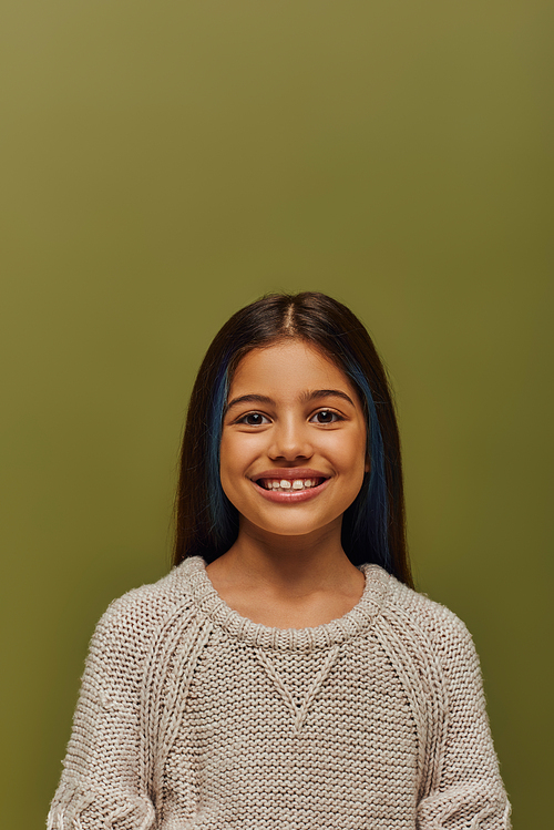 Portrait of positive and stylish brunette preteen girl with dyed hair wearing modern cozy knitted sweater while looking at camera while standing isolated on green, modern preteen fashion