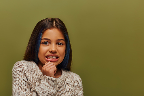 Portrait of disgusted preteen girl with colored hair wearing cozy knitted sweater and looking at camera while standing and posing isolated on green, modern preteen fashion